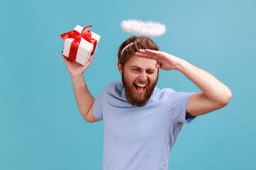 Portrait of positive optimistic bearded angelic man with holy nimbus holding wrapped gift box and...