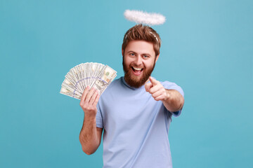Portrait of satisfied bearded angelic man with holy nimbus over head showing fan of money, holding...