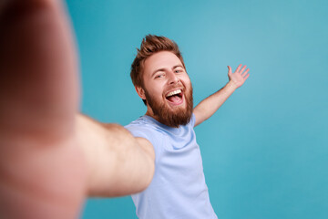 welcome. Portrait of excited satisfied man blogger POV, point of view of photo, laughing happily, and welcoming raised arm, expressing happiness. Indoor studio shot isolated on blue background.