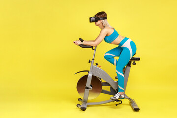 Fototapeta na wymiar Side view portrait of slim woman riding exercise bike with virtual reality headset, doing cardio workout, wearing blue sportswear. Indoor studio shot isolated on yellow background.