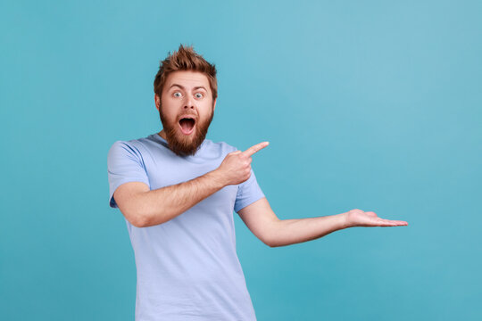 Wow, product advertisement. Portrait of amazed bearded man presenting copy space on palm, showing empty place for commercial text or goods. Indoor studio shot isolated on blue background.