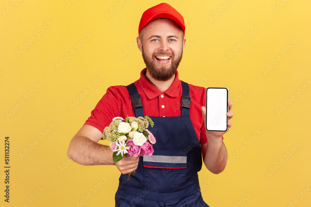 Wall mural Smiling positive man courier standing with flowers in hands and showing cell phone with empty screen for advertisement, looking at camera. Indoor studio shot isolated on yellow background.