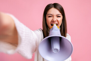 Excited brunette female screaming loud in megaphone, making point of view photo from presentation, wearing white casual style sweater. Indoor studio shot isolated on pink background.