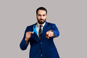 Serious confident bearded man holding blue ribbon, symbol of Colorectal cancer awareness, pointing finger ant camera, wearing official style suit. Indoor studio shot isolated on gray background.