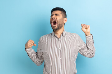 Portrait of young attractive businessman with beard yawning and raising hands up, feeling fatigued of overtime work, wearing striped shirt. Indoor studio shot isolated on blue background.