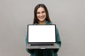 Satisfied delightful woman standing showing laptop with white empty screen for promotion, looking at camera with smile, wearing casual style jacket. Indoor studio shot isolated on gray background.