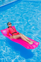 Happy young woman in sunglasses and pink bikini floating on inflatable mattress by the pool. Summer vacation concept. Travel by sea
