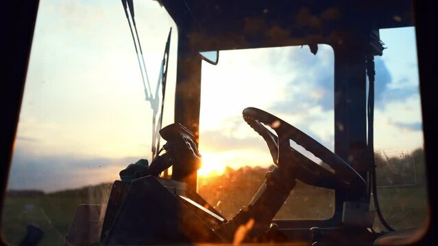 Side view through window of tractor driver cab with steering. Car is standing on field against sunset. Management agriculture with help of equipment. Simplification of production process on farm.