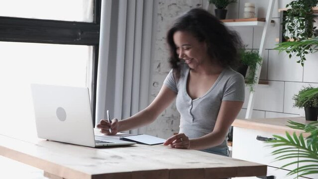 African American woman sits indoor and using app on laptop for video call to friends, making notes in a notebook. Young female employee in casual wear talking online via video connection waving hands