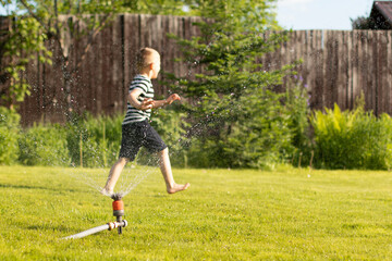 Fototapeta premium A little wet boy runs barefoot on the lawn next to the sprinkler. The concept of a happy, carefree childhood and holidays