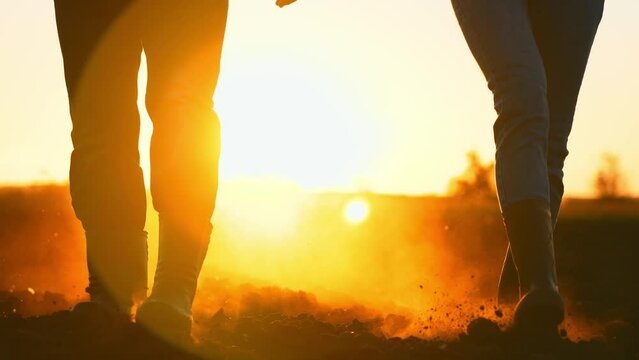 Close up people feet are walking on black earth soil on agricultural land in rubber boots in nature on field in summer at sunset and dust rises in sun. Couple of rural farmers in family business.