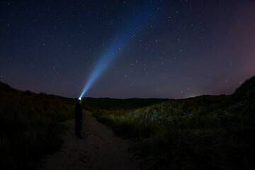 Adventurous man looking up at the stars on a beach path at night with his headlamp shinning up at...