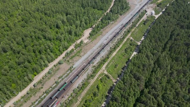 Aerial View above close paralel of the Cargo Long Train in taiga, mountains near to dirt road with tracks in sunny summer afternoon