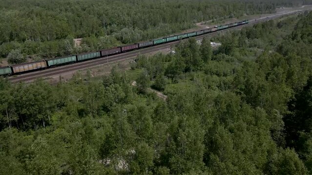 Aerial View parallel above close of the Cargo Long Train in taiga, mountains near to dirt road with tracks in sunny summer afternoon