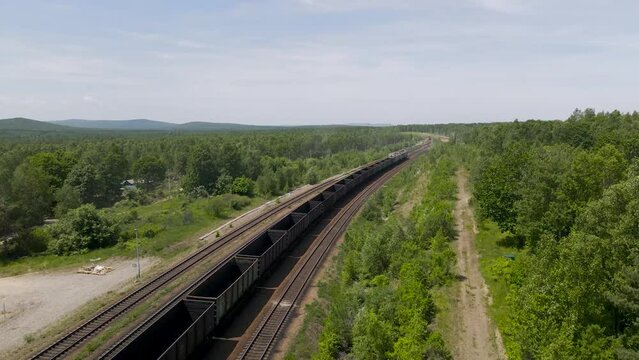 Aerial View parallel above of the Cargo Long Train in taiga, mountains near to dirt road with tracks in sunny summer day
