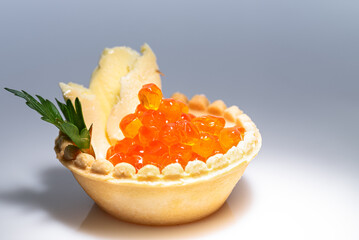 Red caviar on tartlet on beige oil with basil leaf on gray background is illuminated by white light.