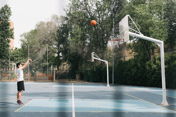 Dark-haired boy shooting a street basketball free throw. Image from behind a fence of a lone young...