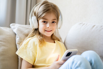 Adorable blonde girl wearing wireless headphones, watching funny video in social network on smartphone or playing online mobile games.