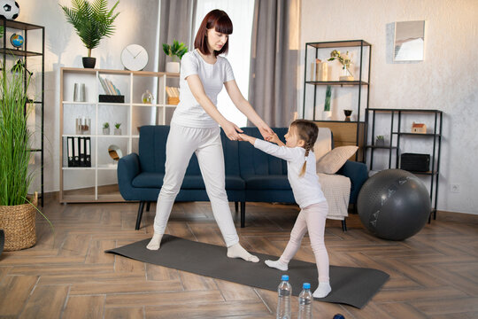Full length portrait of woman wearing white t shirt and leggins doing sport exercises at home with her daughter, standing on mat, stretching body, training together.