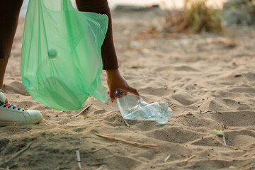 Cropped photo of African woman collecting spilled trash garbage from sand on beach in green plastic bag. Womans hand cleaning up used plastic bottles. Ecology, environmental conservation, pollution. 
