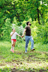 Naklejka na ściany i meble Portrait of smiling family walking pacing in forest around green trees, having fun. Little cheerful daughter holding hand of middle-aged happy bearded man father. Love, summer activities. Vertical.