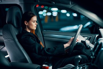 a horizontal photo from the side, at night, of a woman sitting behind the wheel in a black shirt,...