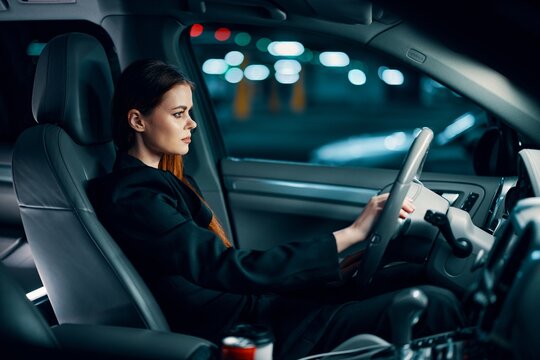 A Horizontal Photo From The Side, At Night, Of A Woman Sitting Behind The Wheel In A Black Shirt, Wearing A Seat Belt, Looking At The Road. Safe Driving Topics