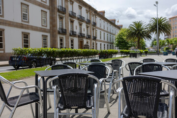 An empty outdoor cafe on a sunny day with wicker chairs and black tables. In the background is a beautiful residential building, palm trees, an artisanal hedge. Siesta. Hot. Quarantine. The absence of