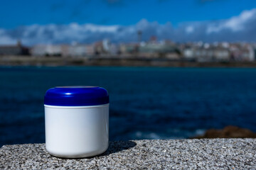 Obraz na płótnie Canvas A white jar of cream with a blue lid stands on a stone surface. In the background is the ocean. Blue bright sky. Blue Ocean. A sunny day. Useful properties. Ecological compatibility. Naturalness.