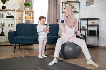 Happy Muslim family doing sports in morning at home. Beautiful Arab woman sitting on fitball and doing exercises with dumbbells while her little daughter sitting next to her with a bottle of water.