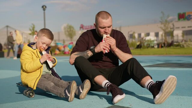 Father and son spend time together eating food sitting on asphalt in park. Happy father spends time with his young son in park, eating fast food in summer in sunny park.