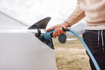 Obraz premium Male hand putting a charger in electric car socket, in the background visible mountain nature