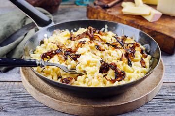 Traditional Swiss Käsespätzle with roasted onions served as close-up in a iron frying pan on a rustic wooden board at a mountain chalet