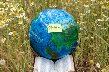 Earth globe with a handwritten word peace on a note and open Holy Bible Book in a field with green...