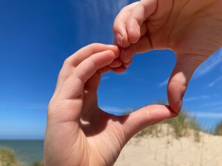 Kinderhände formen ein Herz am Nordseestrand mit Dünen, blauem Himmel, Sand und Meer