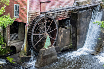 A historic wooden mill and operating water wheel and a dam on a river.
