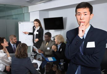 Thoughtful serious Korean man standing at office on background with working colleagues