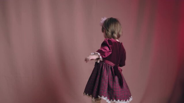 Cute little girl in an vintage burgundy dress is playing with a rattle toy. The concept of children theater productions and developing acting circles. Pink background