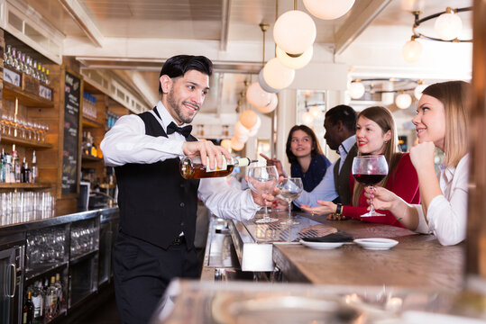 Smiling bartender pouring beverages and talking to cheerful visitors in bar.