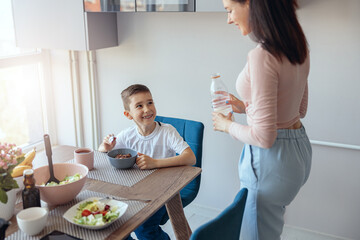 Small Caucaian boy sitting at kitchen table with bowl of cereals, mother with milk.