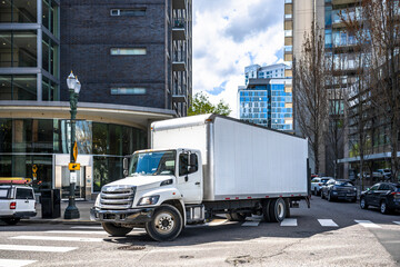 Big rig day cab white semi truck with long box trailer making local commercial delivery at urban city with multilevel residential apartments buildings turning on the city street with crossroad - Powered by Adobe