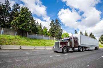 Fototapeta na wymiar Classic burgundy big rig semi truck tractor transporting cargo in dry van semi trailer driving on the wide highway road with clouds sky