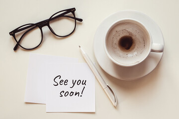 See you soon note, empty cup with coffee leftovers, eyeglasses and pen on white desk
