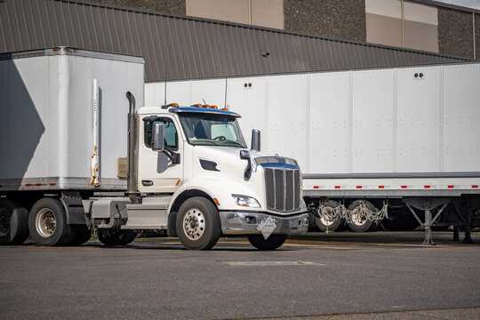Day cab white big rig semi truck with dry van semi trailer unloading cargo standing in warehouse dock