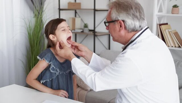 Child healthcare. Pediatrician checkup. Kid flu diagnosis. Middle-aged male doctor examining sick small girl patient sore throat at modern interior.