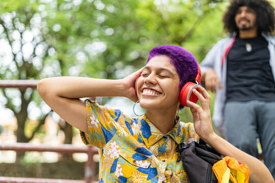Mujer latina bisexual con el cabello corto y de color morado, disfrutando de música con unos auriculares rojos al aire libre con una gran sonrisa de felicidad y su amigo con cabello afro viendo atrás.