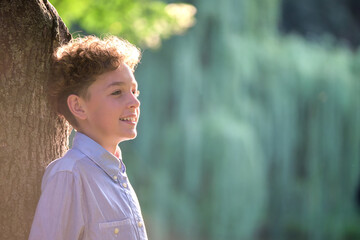 Fototapeta premium Young happy child boy relaxing in summer park leaning on tree trunk. Child wellbeing concept