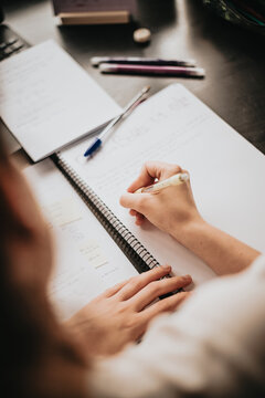 Top view of the back of a young woman studying and working on his home desk, doing homework during university, preparing for exam with textbook and taking notes, selective focus on the pencil.