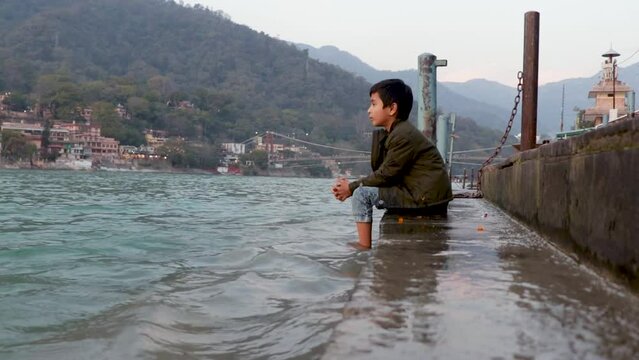 isolated young kid sitting at the shore of holy ganges river bank from flat angle video is taken at ganga river bank rishikesh uttrakhand india.