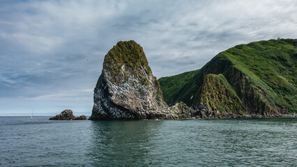 Fototapeta na wymiar A picturesque island in the Pacific Ocean. Seabirds nest on steep rocky slopes. A green hill against a background of blue sky and clouds. Kamchatka. Starichkov Island